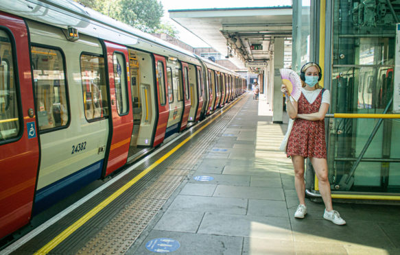 A passenger wearing a facemask standing on the London Underground platform using a fan to cool down from the heat and humid conditions during the August heatwave.